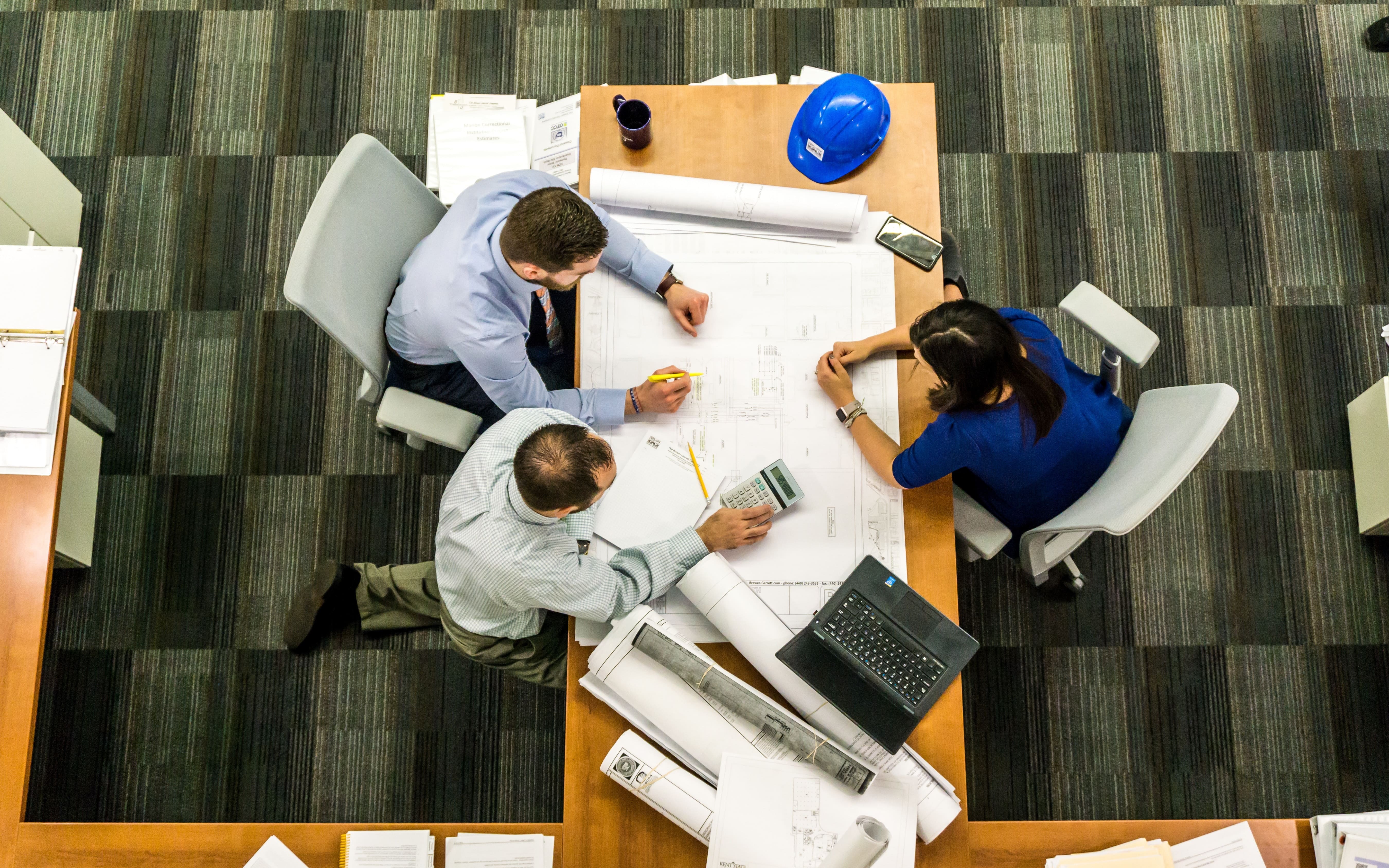 Three people sit at a table with blue prints. A blue hard hat can be seen. 