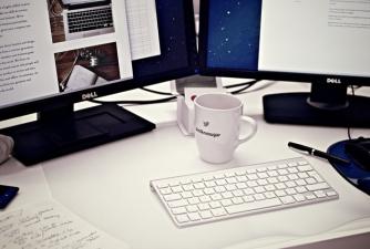 A mug and keyboard in front of two screens